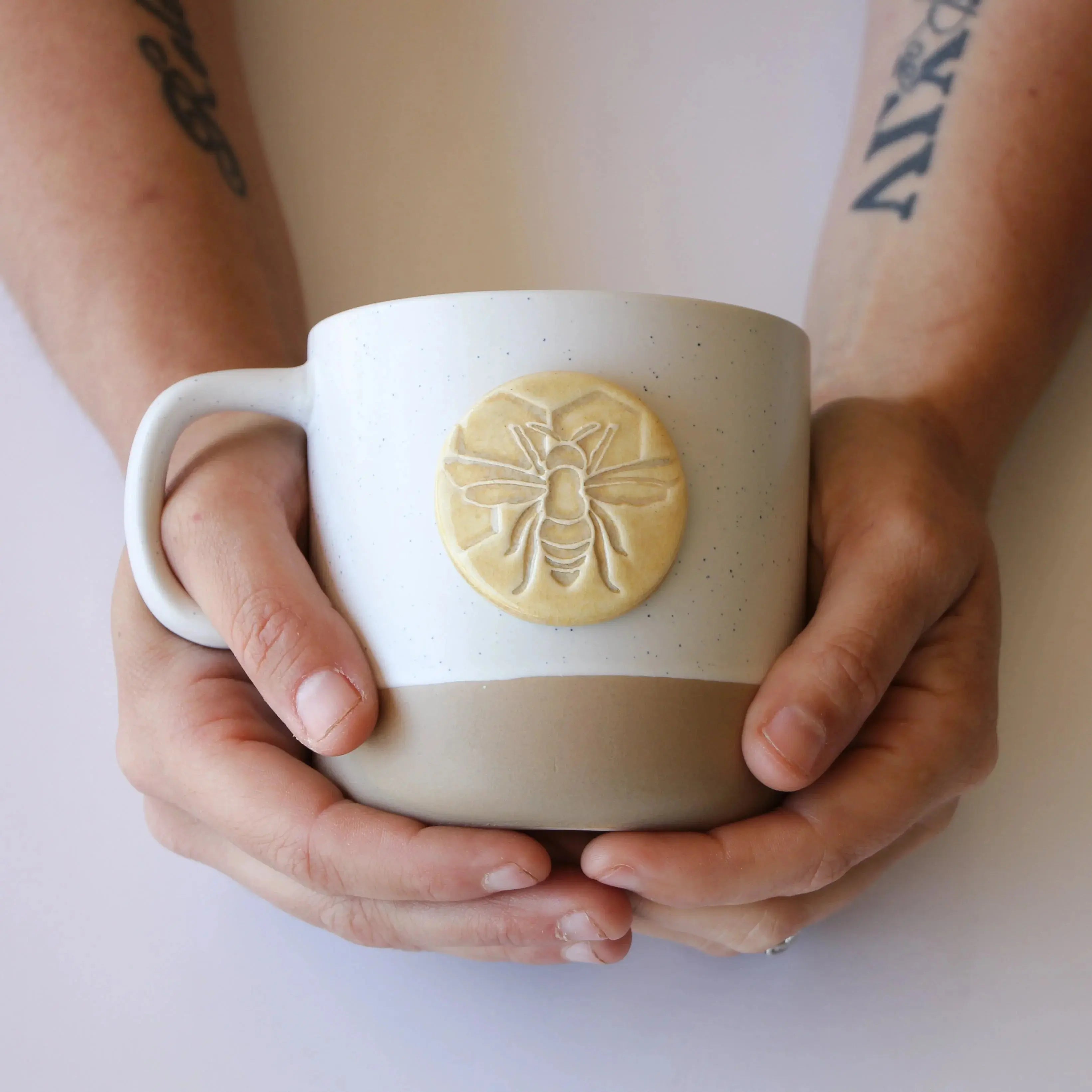 Hands holding a speckled ceramic mug with a bee emblem, cozy farmhouse decor