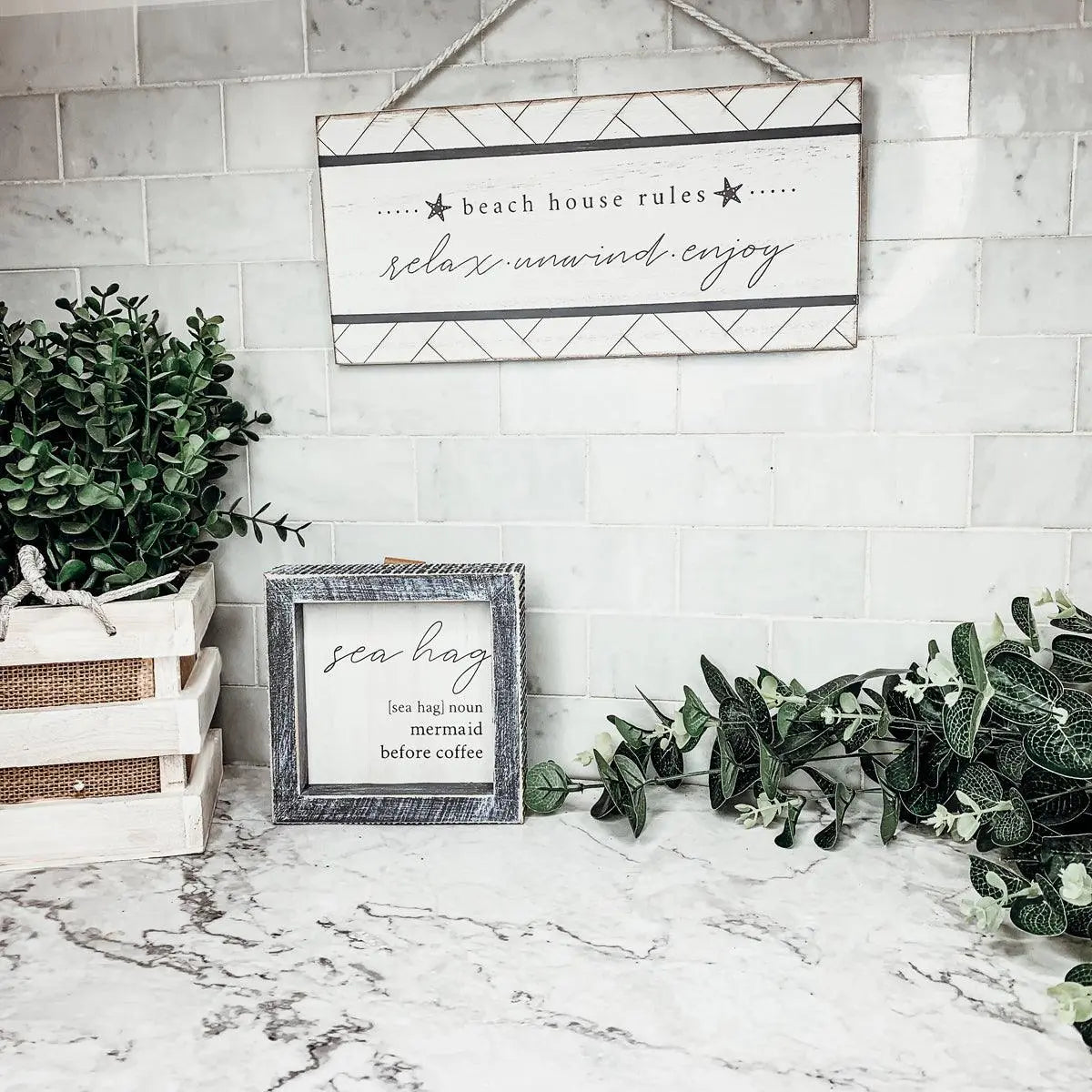 Beach house rules sign with relax unwind enjoy text, rustic frame, greenery, and marble countertop decor