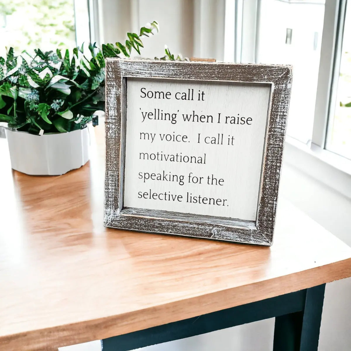 Framed motivational quote on a wooden table with a plant in the background