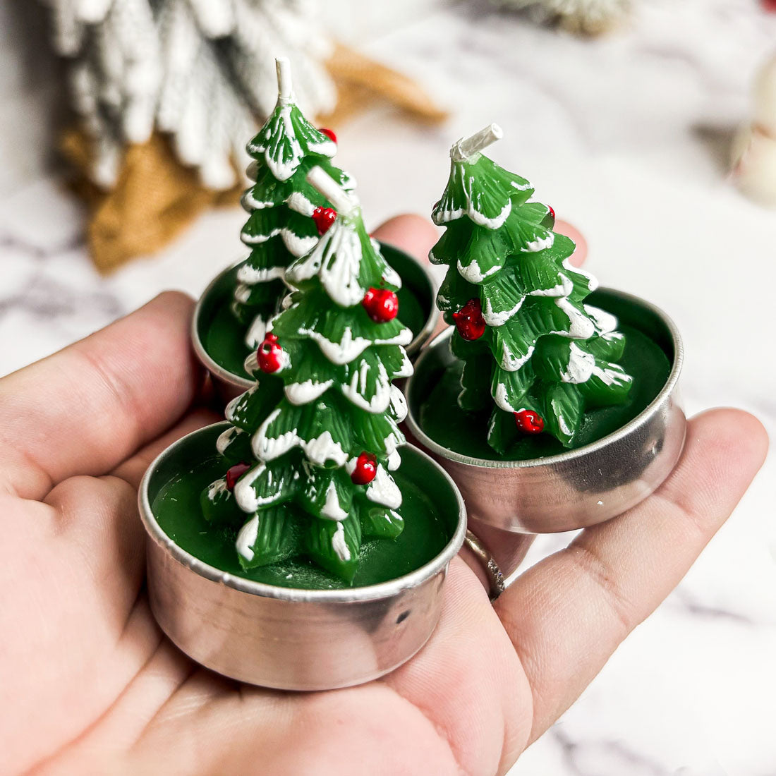 Hand holding four Christmas tree-shaped candles with a snowy top and red berries.