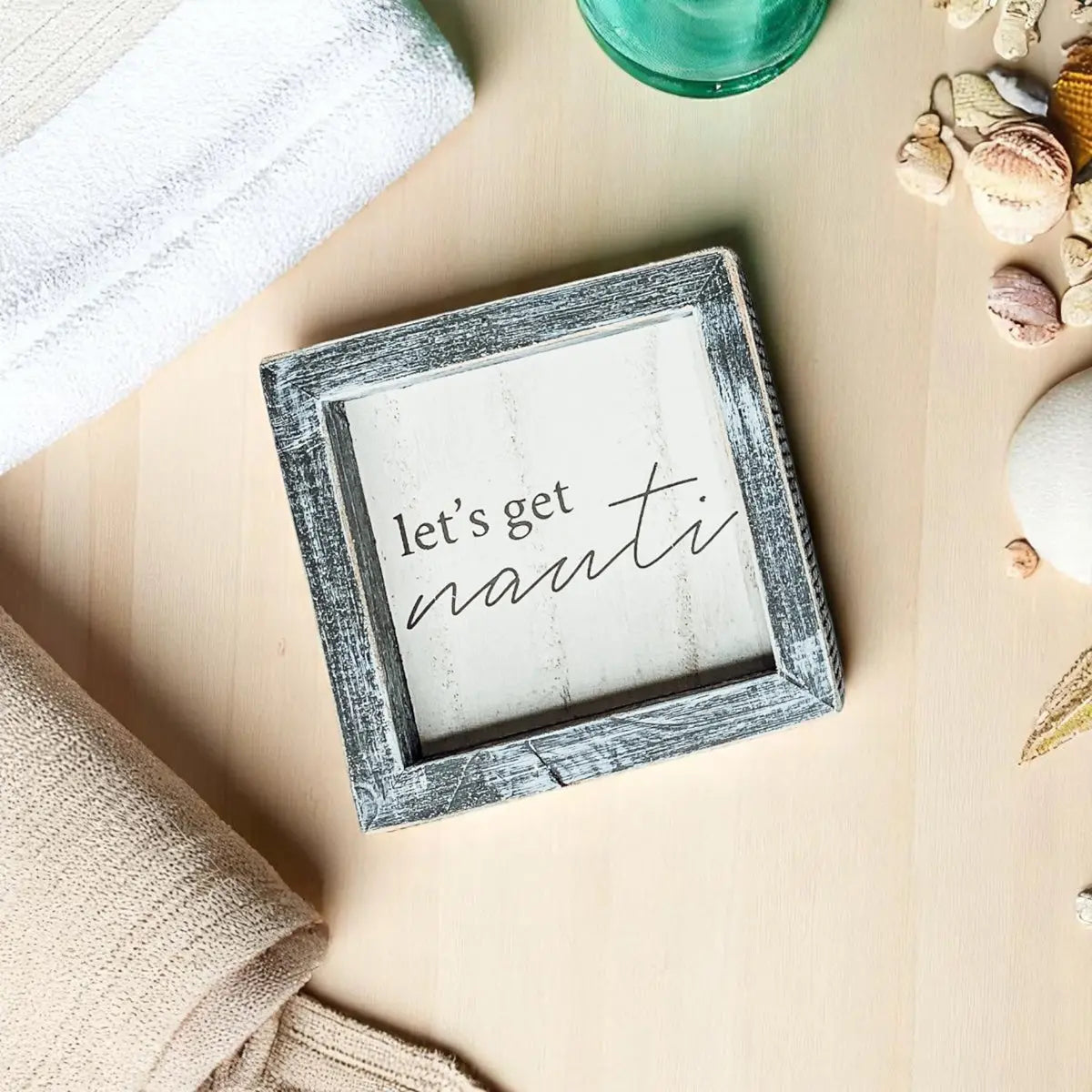 Decorative sign with 'let's get nauti' text on a beach-themed setting with shells and towel.