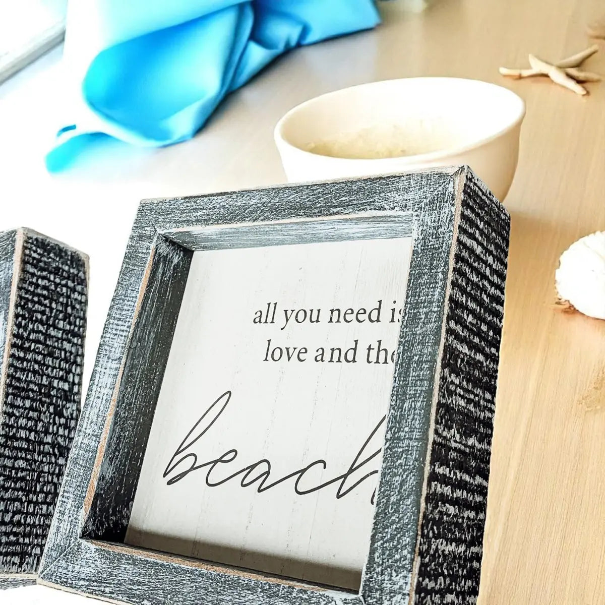Decorative sign with 'all you need is love and the beach' text in a textured frame on a wooden surface.