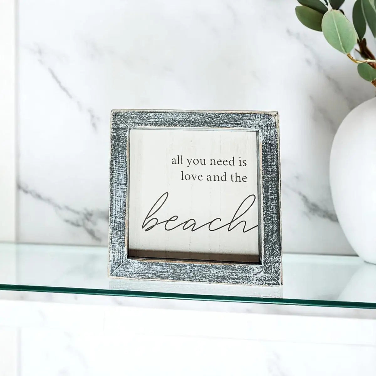 Framed sign with 'all you need is love and the beach' text on a marble surface.