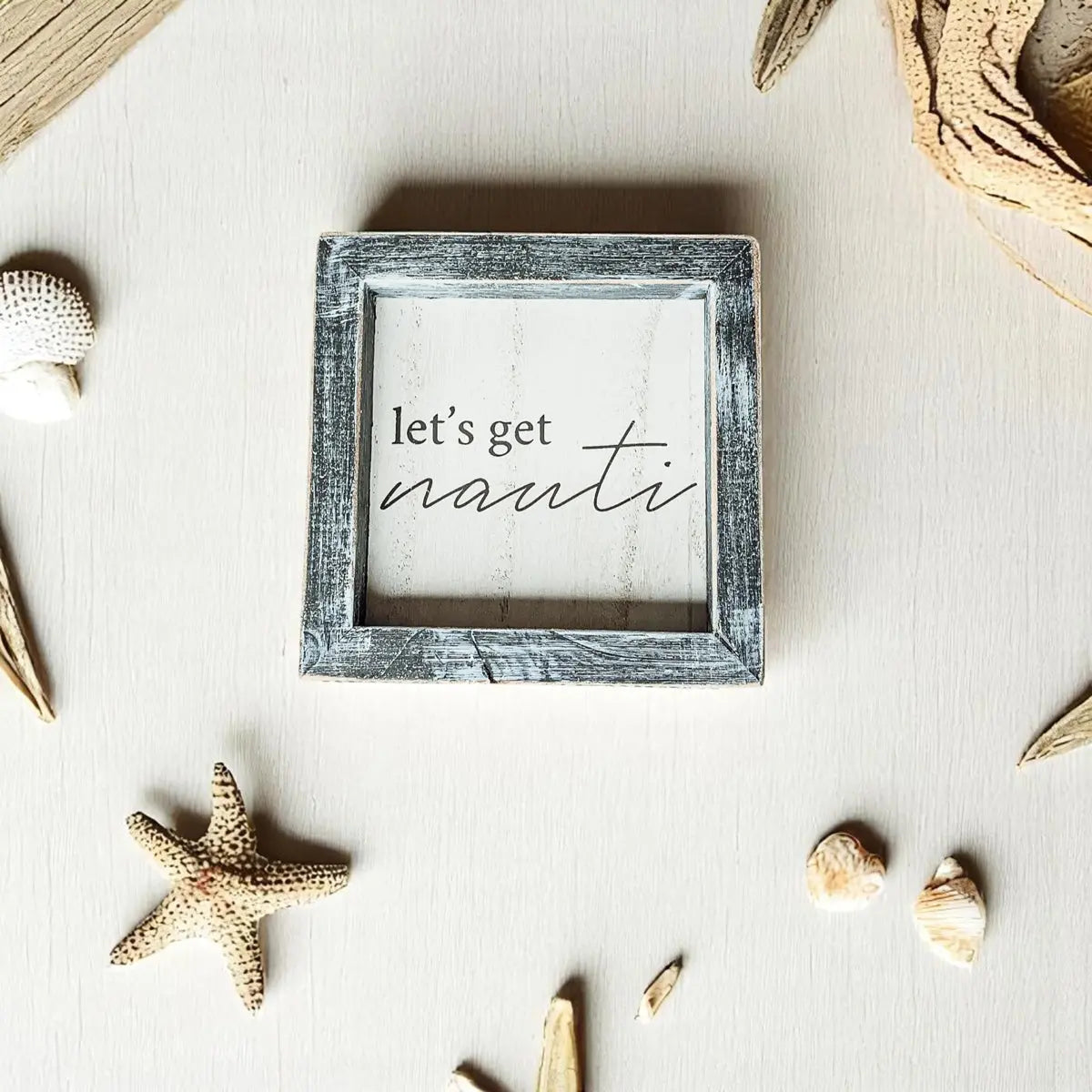 Decorative sign with 'let's get nauti' text surrounded by beach-themed items on a light surface.