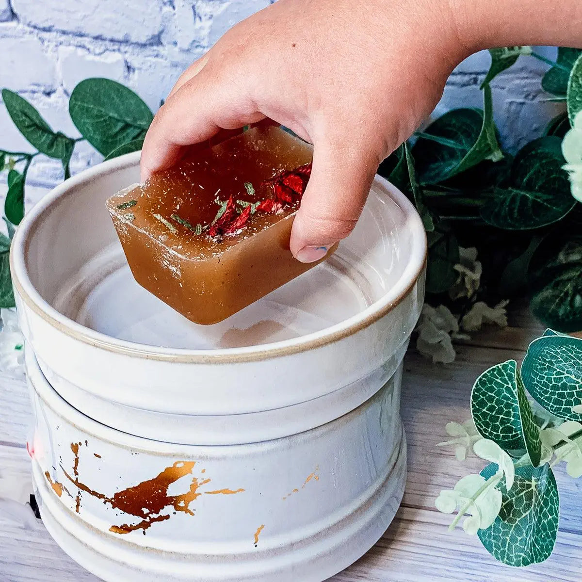 Hand holding a piece of brown soap over a white container with greenery in the background