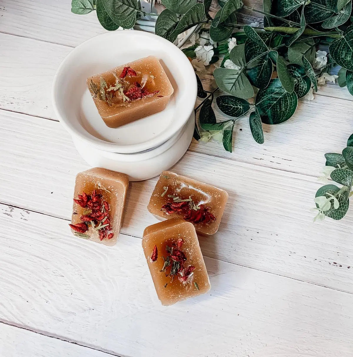 Brown soap bars with red and green herbs on a white wooden surface with green leaves.