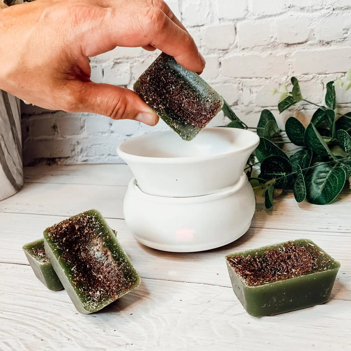 Hand holding a green soap bar over a white ceramic diffuser with plants in the background.