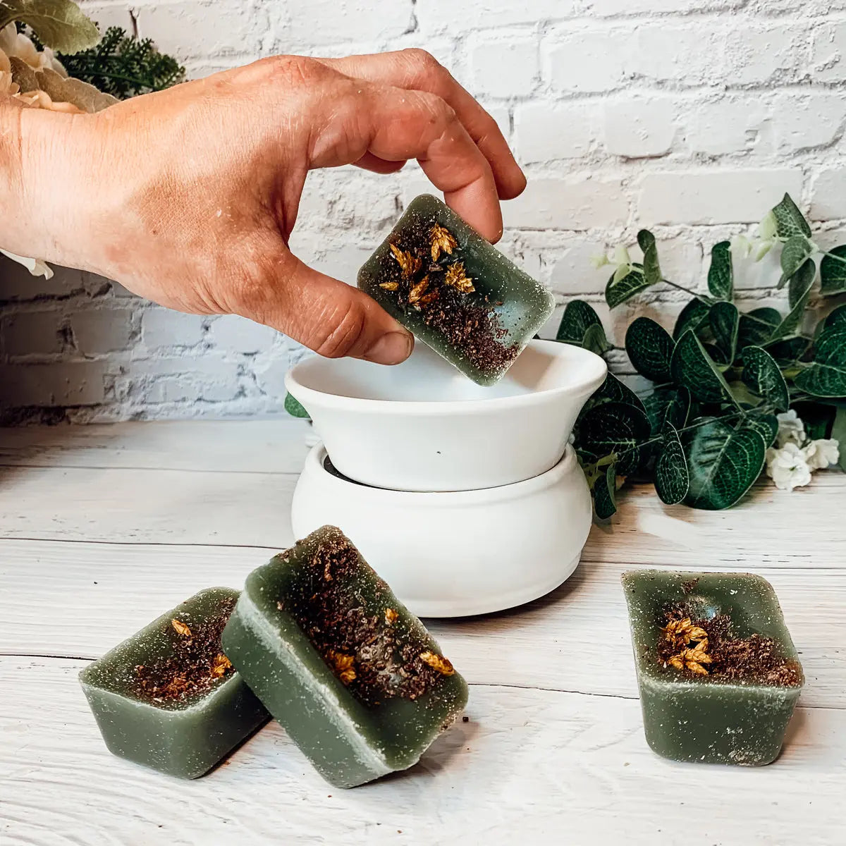 Hand dipping a green wax cube into a white wax warmer with plants in the background.