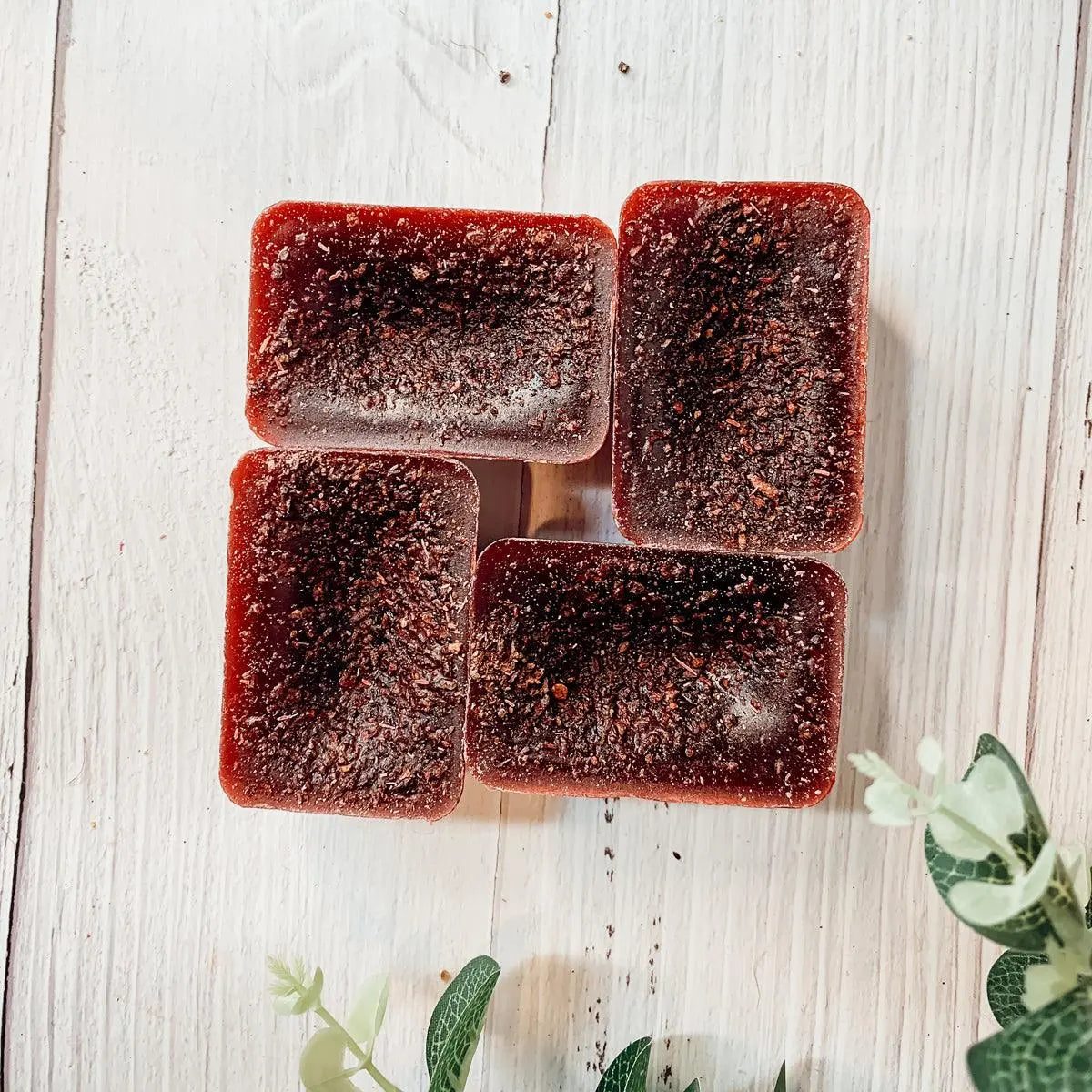 Four rectangular dark red soap bars on a light wooden surface with green leaves in the corner.