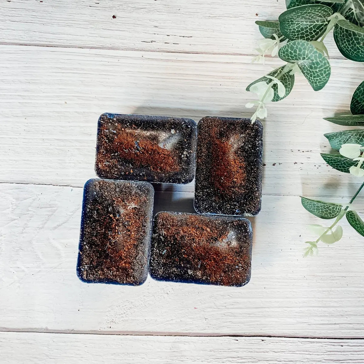 Four rectangular dark-colored soap bars on a light wooden surface with green leaves.