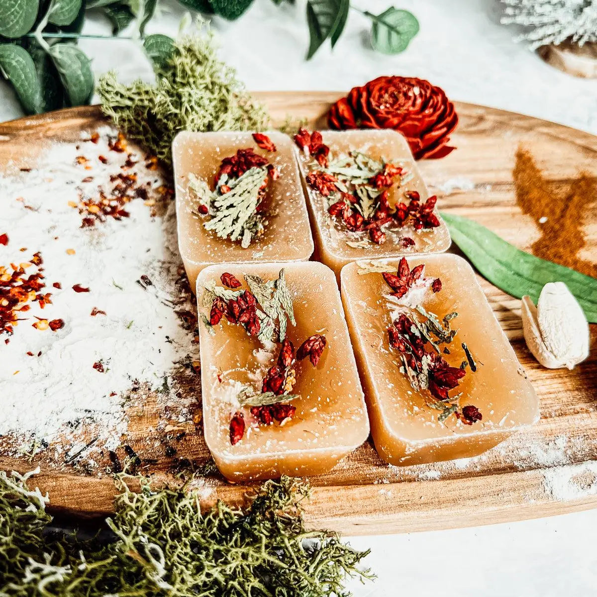 Four square soap bars with natural ingredients on a wooden board with greenery.
