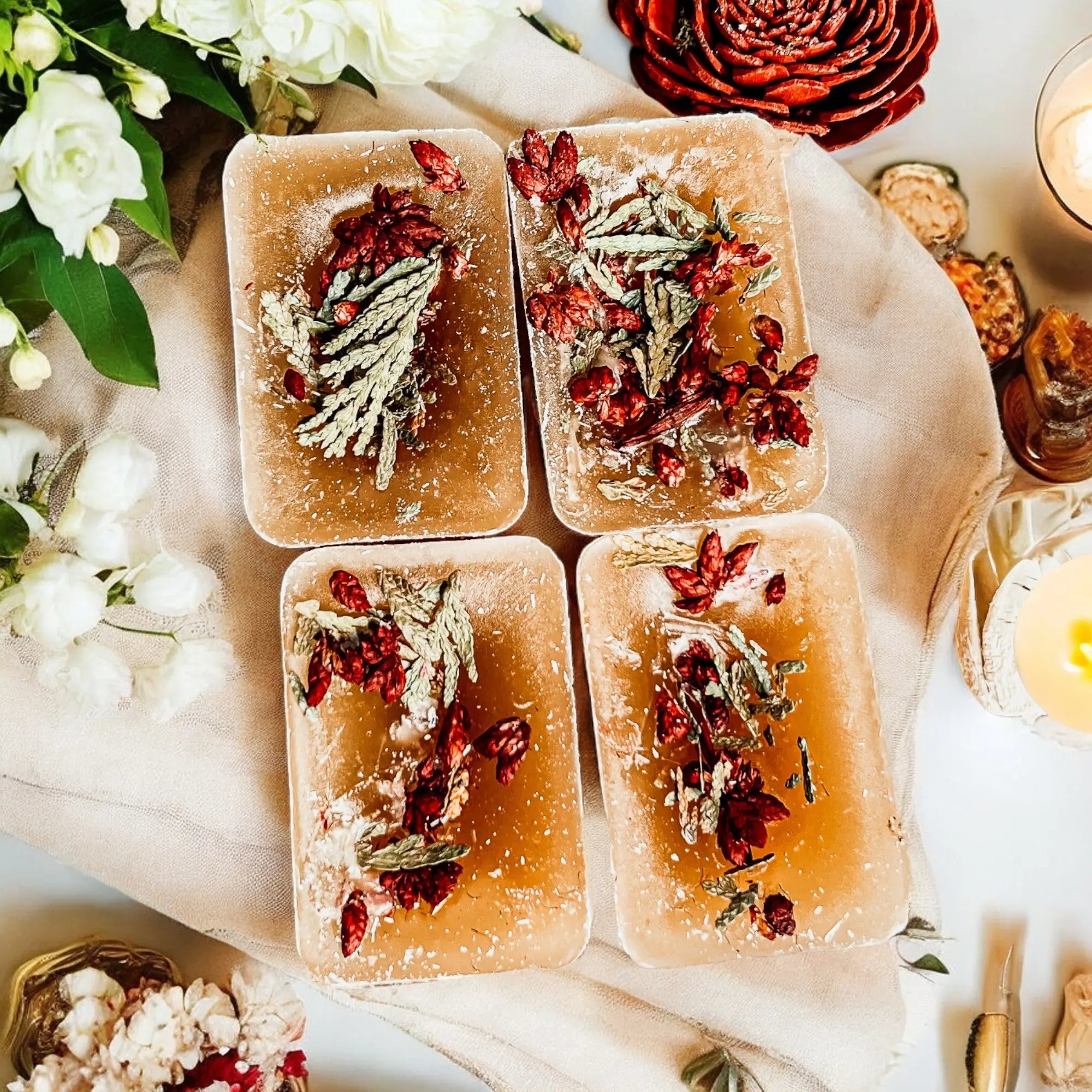 Four rectangular soap bars with dried herbs on a decorative surface with flowers and candles.