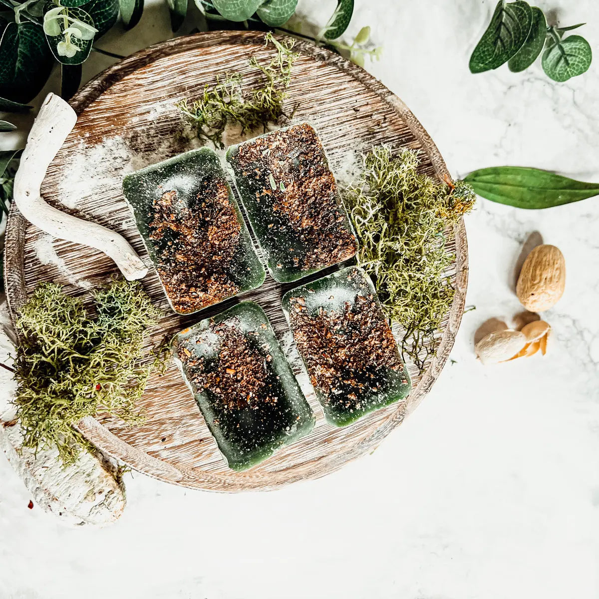 Green soap bars with brown specks on a wooden tray surrounded by greenery
