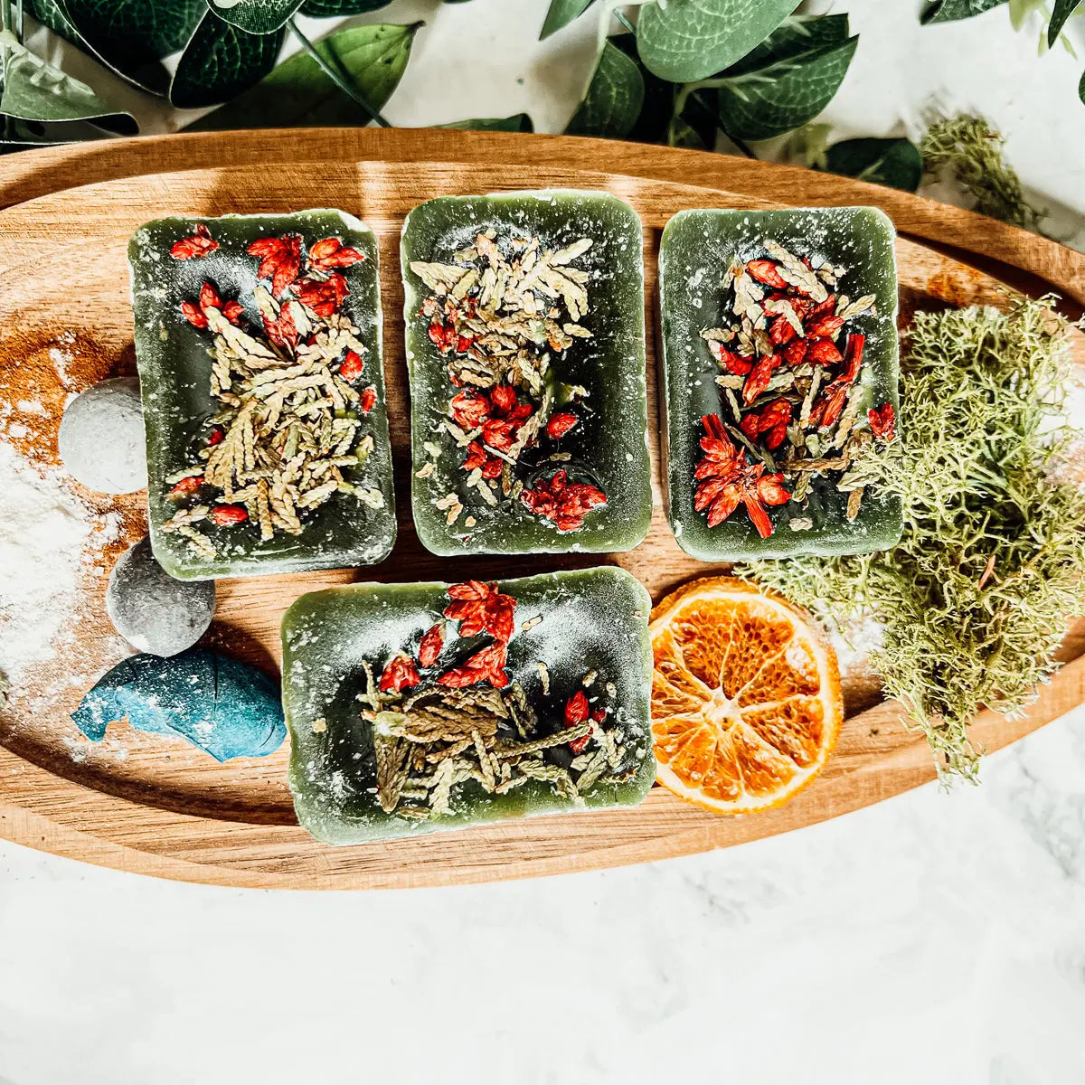 Four square ice cubes with herbs and a dried orange on a wooden tray with greenery in the background.
