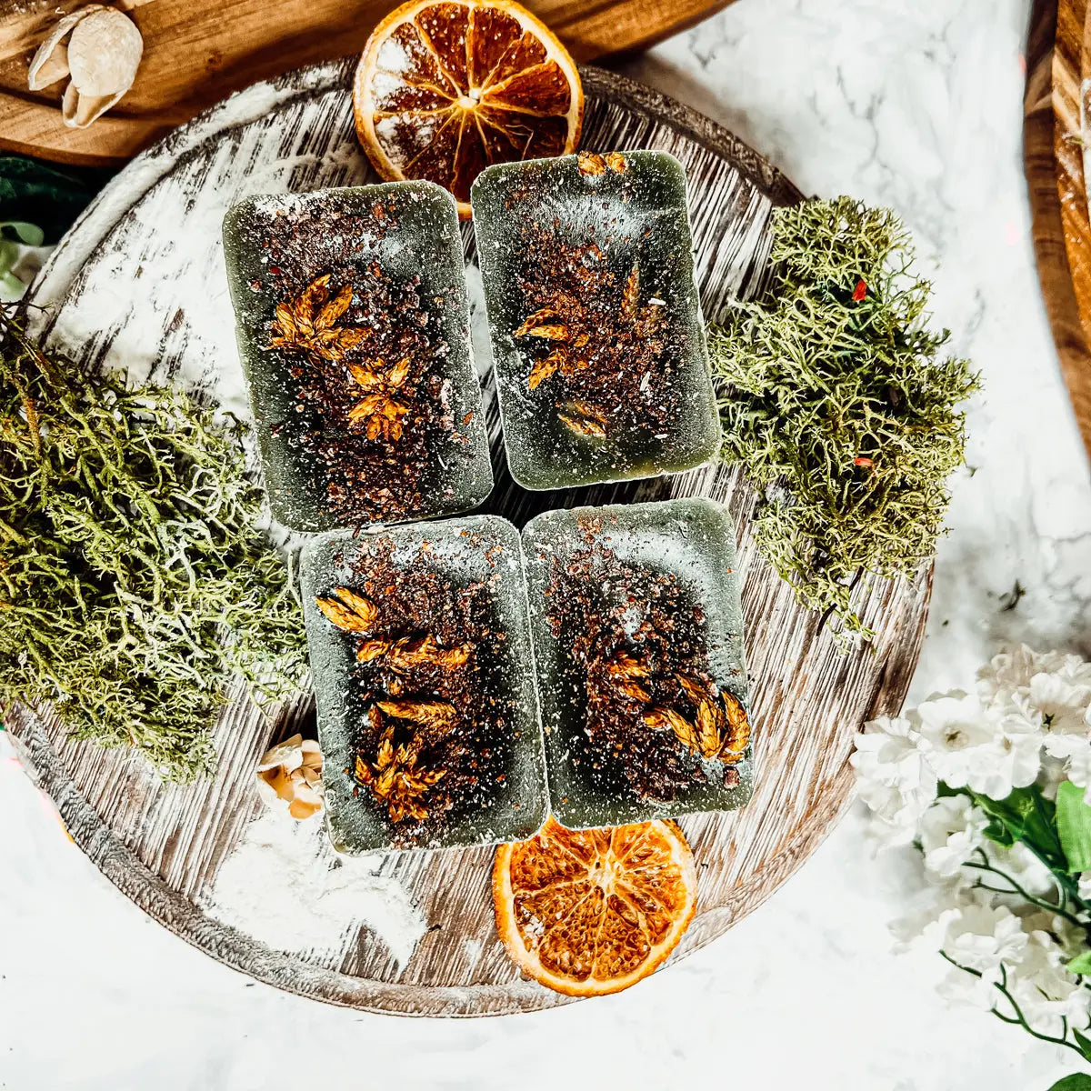 Five dark green soap bars with brownish specks on a wooden board with dried oranges and moss.