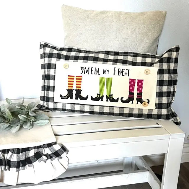 A decorative rectangular throw pillow with a black-and-white gingham border and a cream center panel that reads “SMELL MY FEET” above illustrated witch boot socks in orange, green, and pink. The pillow is styled on a white bench with a beige pillow behind it and a small greenery prop at left.