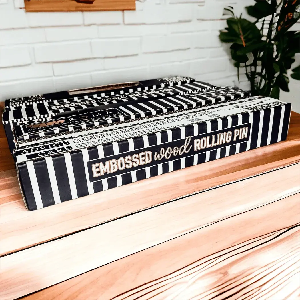 Box of embossed wood rolling pins on a wooden surface with a white brick wall in the background.
