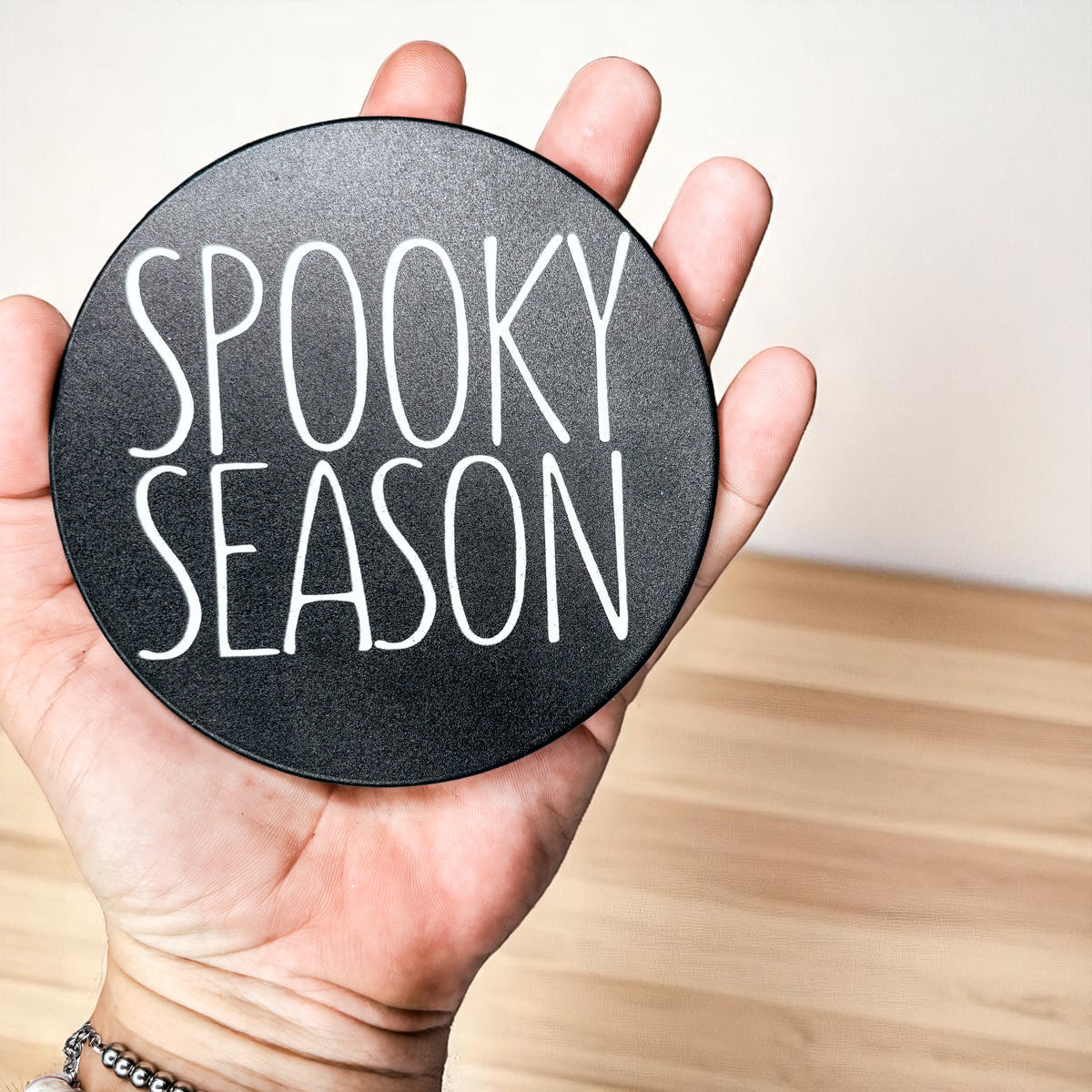 Hand holding a round black sign with 'Spooky Season' text against a neutral background