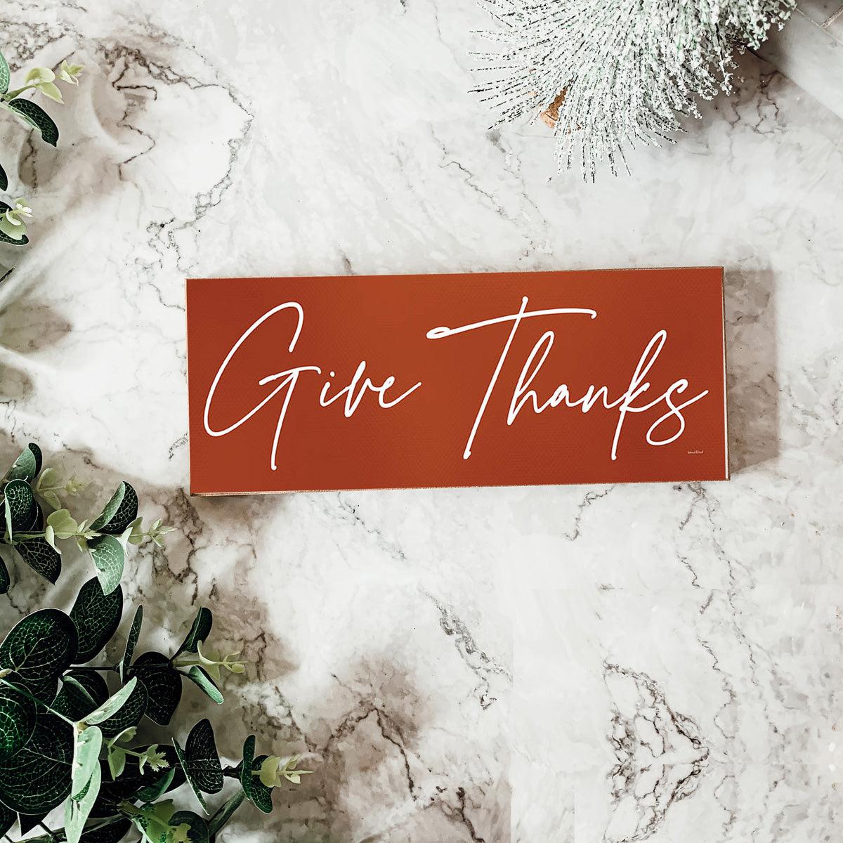 Red sign with 'Give Thanks' on a marble surface with greenery
