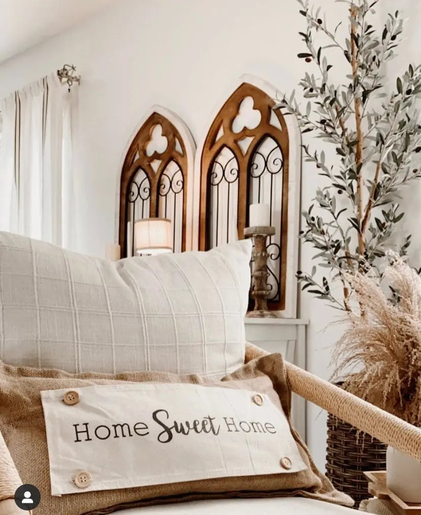 A cozy home interior scene featuring a beige and brown throw pillow with the words 'Home Sweet Home' embroidered on it, styled on a sofa next to a tall potted olive tree and decorative wall panels.