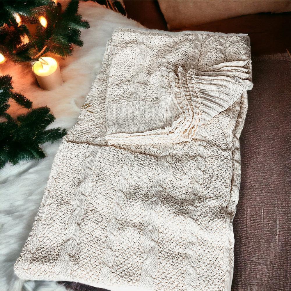 Cozy cream cable knit blanket with fringed edges, styled with holiday greenery and candlelight for warm farmhouse decor.