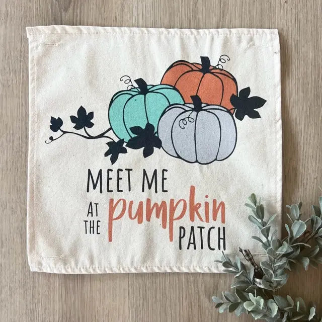 A square cream fabric napkin or small towel laid flat on a light wood surface, printed with three illustrated pumpkins in teal, orange, and pale gray, black leaves, and the phrase “MEET ME AT THE pumpkin PATCH.” A small greenery sprig appears at the lower right corner.
