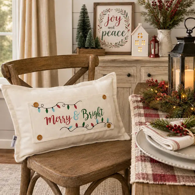 A cream fabric holiday throw pillow with 'Merry & Bright' embroidered in red and green, decorated with colorful string lights and wooden buttons, resting on a rustic wooden chair in a cozy, decorated living room.