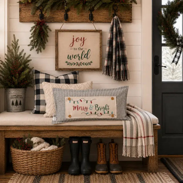 A styled holiday porch scene with a rectangular striped throw pillow featuring a 'Merry & Bright' Christmas string light design, placed on a wooden bench next to a plaid and cream pillow. The setting includes a sign reading 'joy to the world', a plaid scarf, boots, a basket, and a door wreath.