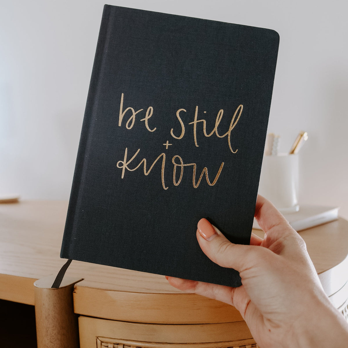 Black notebook with 'be still and know' text held by a hand, blurred background
