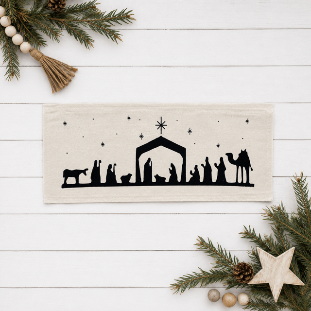 A rectangular cream Pillow Panel featuring a black silhouette of a Christmas nativity scene, displayed on a white wood plank background with festive pine branches, a wooden star, and beads as props.