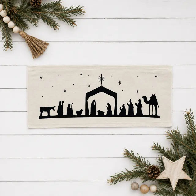 A rectangular cream Pillow Panel featuring a black silhouette of a Christmas nativity scene, displayed on a white wood plank background with festive pine branches, a wooden star, and beads as props.