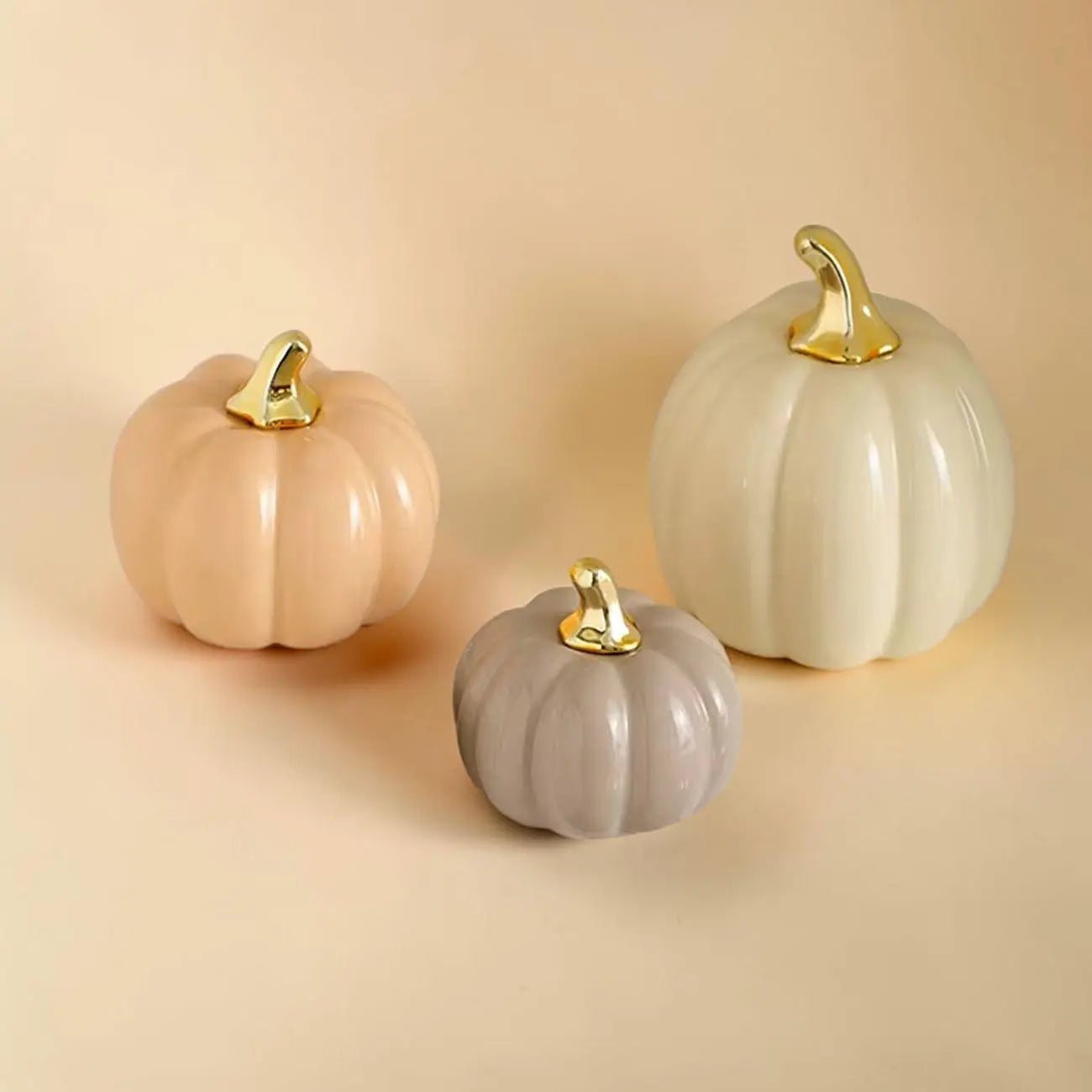 Three decorative pumpkins in beige, gray, and white with gold stems on a beige background