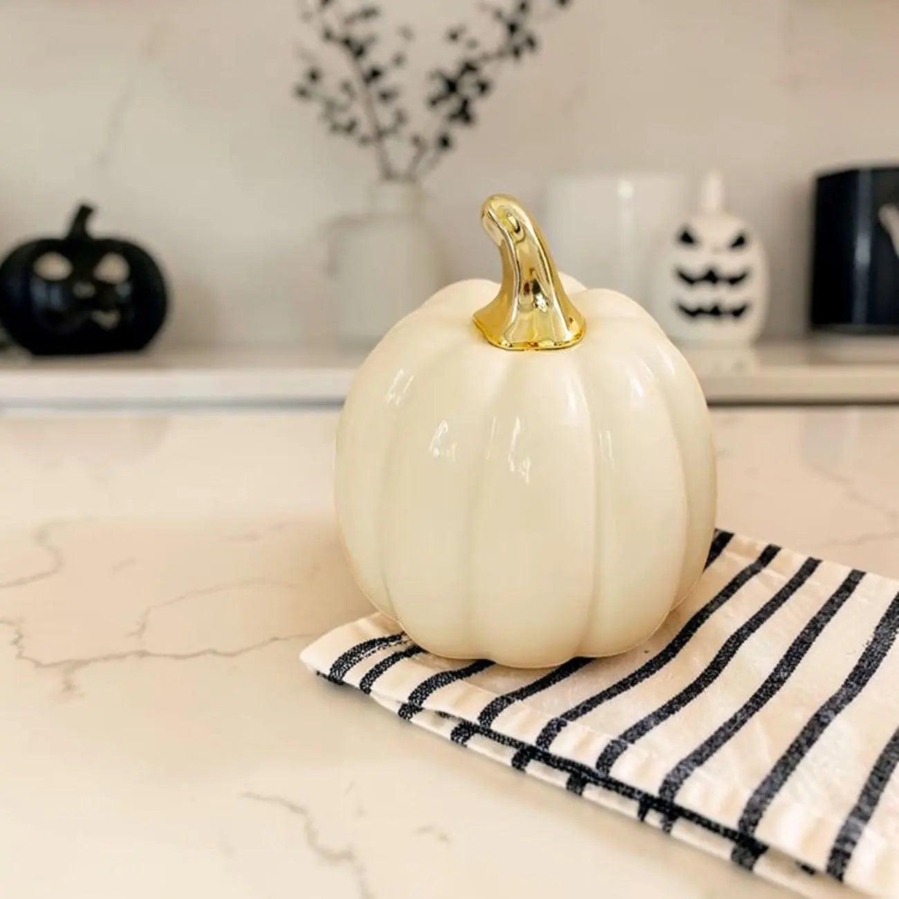 White pumpkin with a gold stem on a marble surface with Halloween decorations in the background.