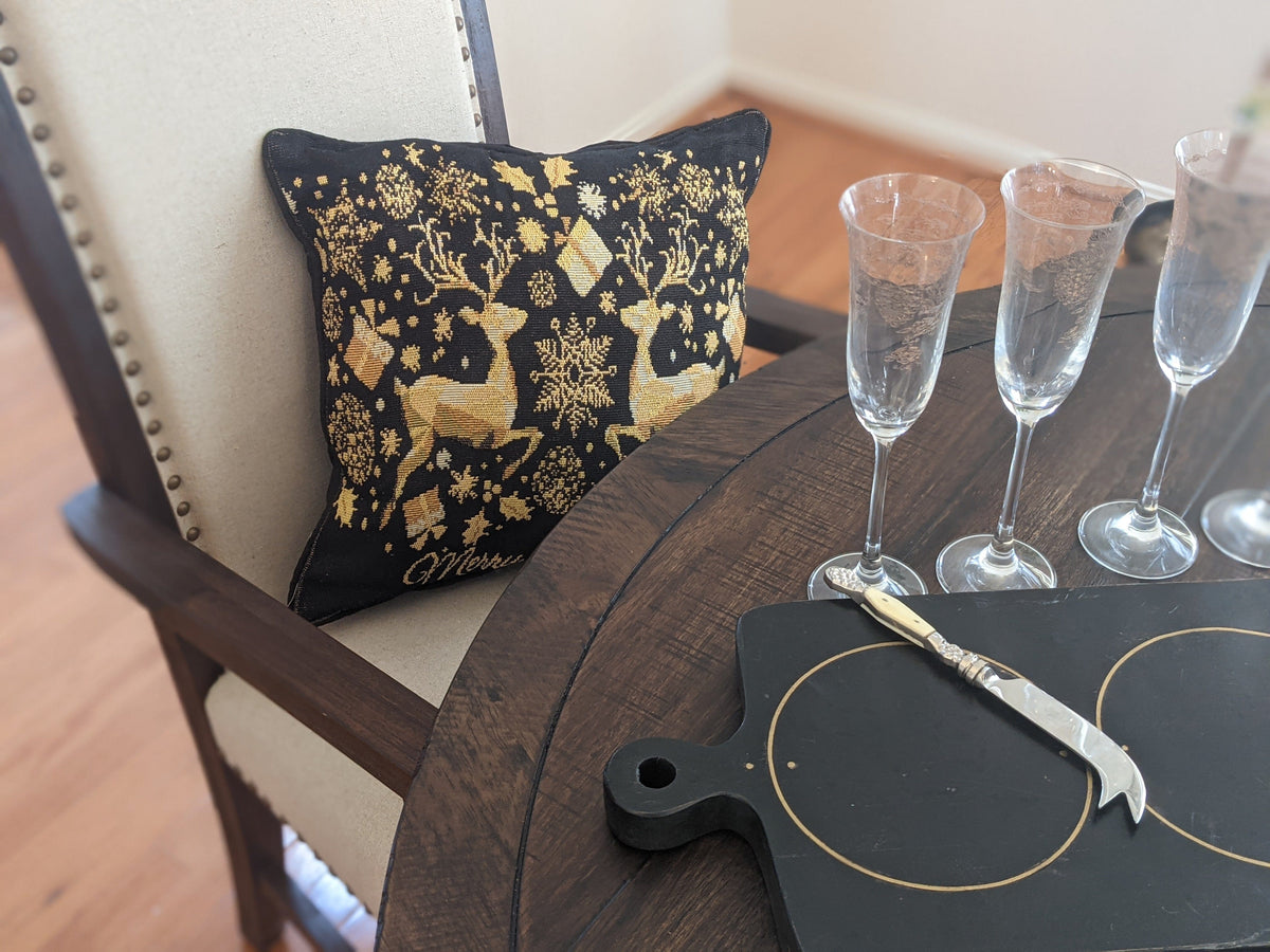 Decorative pillow with gold reindeer design on a chair next to a table with champagne glasses and a cutting board.