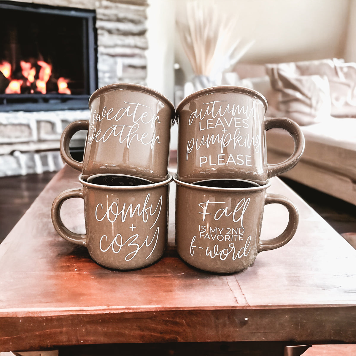 Four brown mugs with autumn-themed text on a wooden table in front of a fireplace.