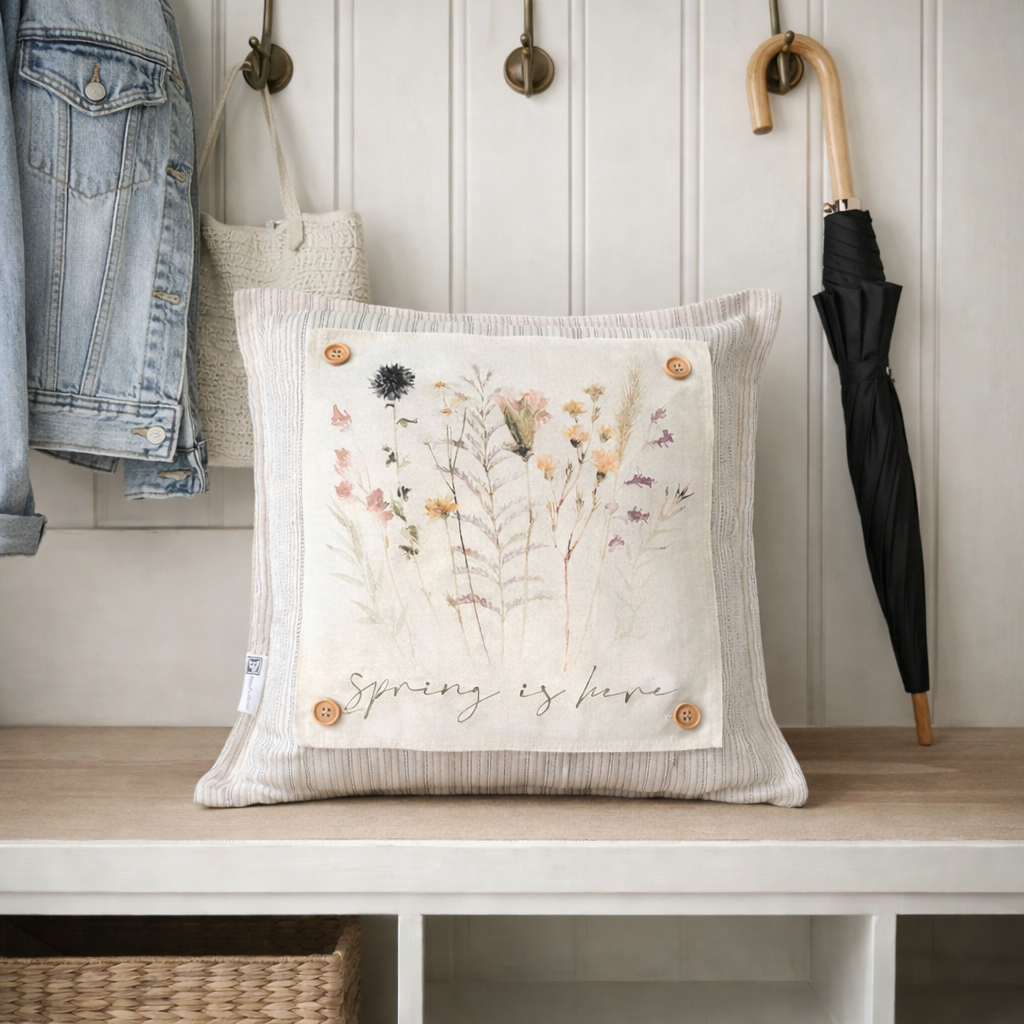 A square decorative throw pillow in a ribbed cream fabric with a central printed panel showing wildflowers and the words 'Spring is here,' styled on a wooden bench in a home interior with a jacket, bag, and umbrella nearby.