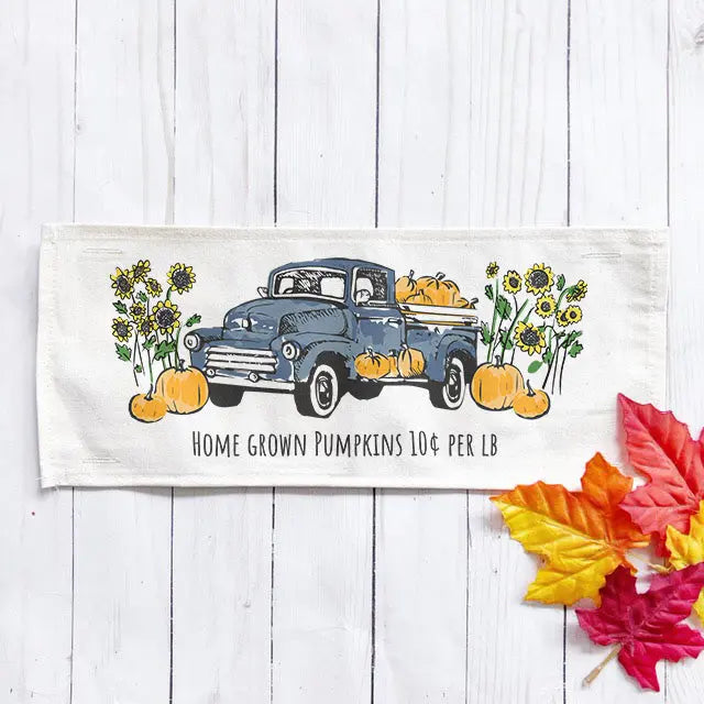 An off-white fabric kitchen towel or placemat featuring a vintage blue pickup truck loaded with pumpkins, surrounded by sunflowers and the text 'HOME GROWN PUMPKINS 10¢ PER LB'. The item is displayed on a white wood plank background with decorative autumn leaves in the lower right corner.