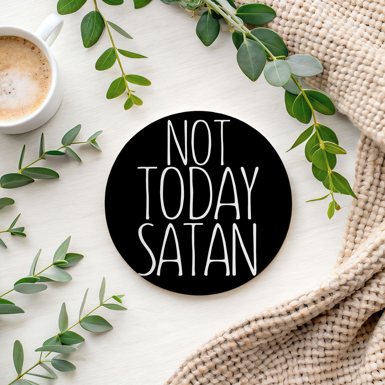 Round black sign with 'Not Today Satan' text on a white surface with green leaves and a cup of coffee.