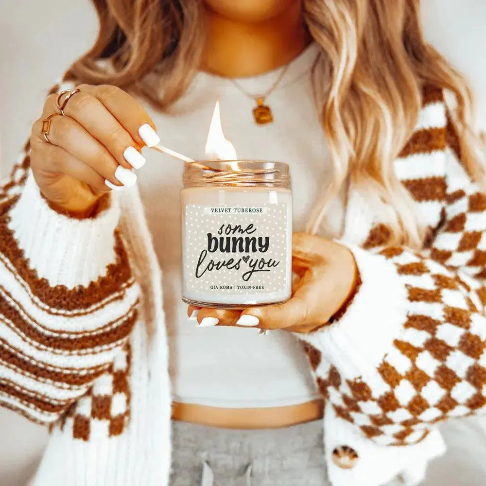 Person lighting a candle labeled 'some bunny loves you' with a cozy sweater and background.