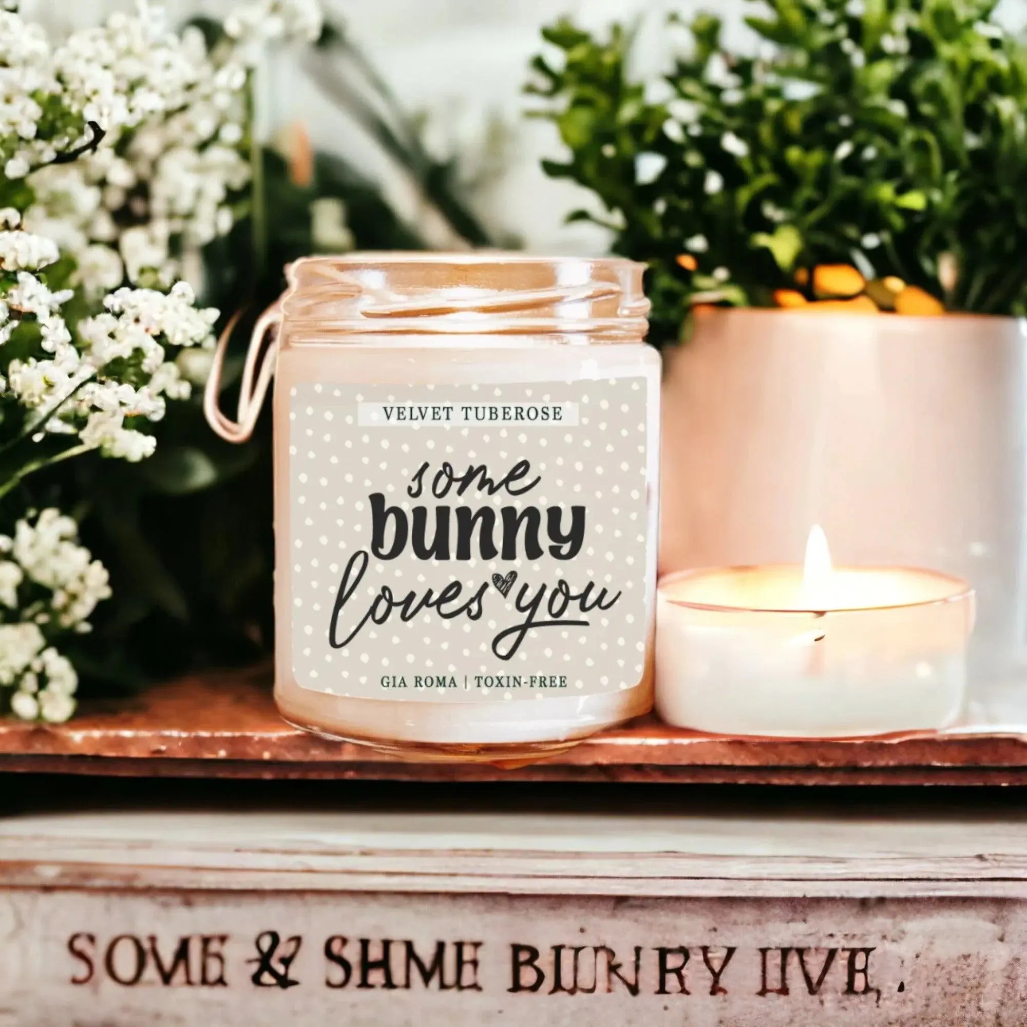 Candle labeled 'Some Bunny Loves You' with a small candle next to it on a wooden surface with plants in the background.