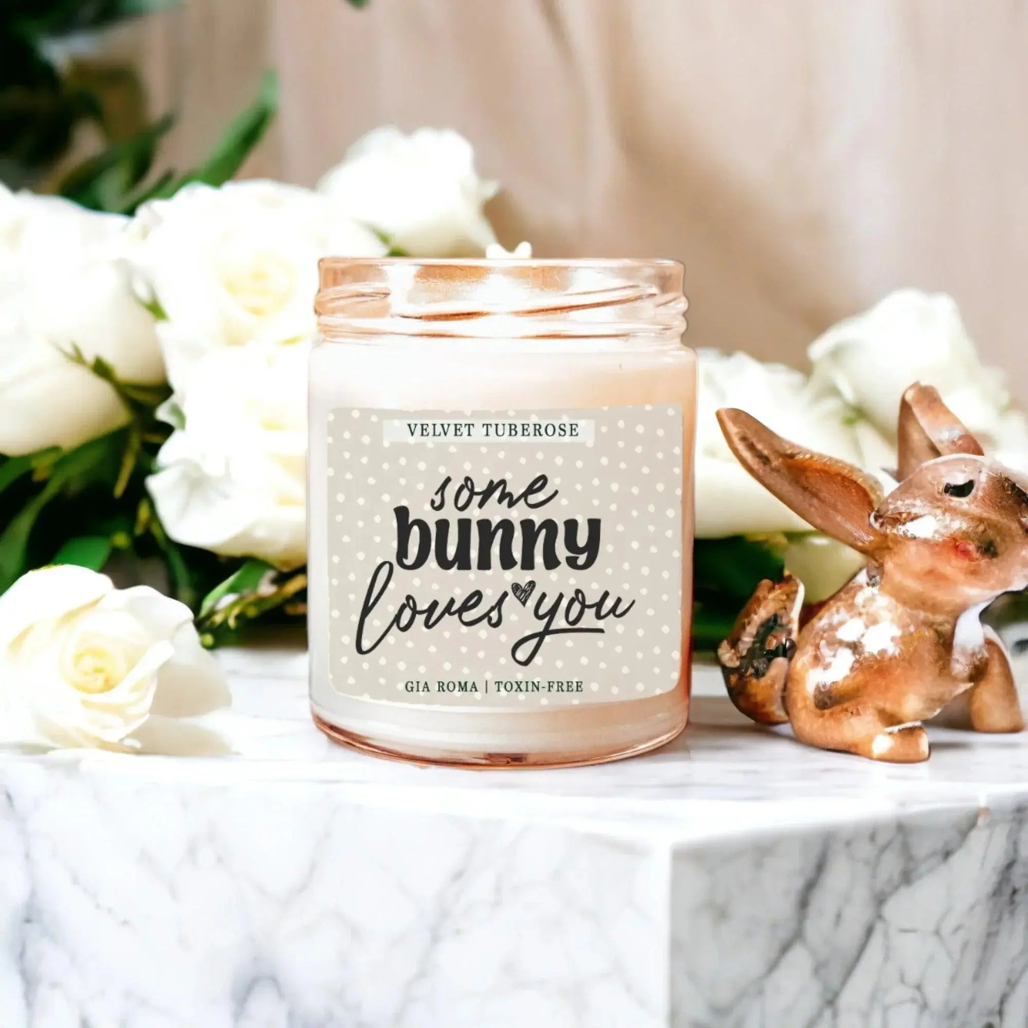 Candle with 'some bunny loves you' text on a marble surface with flowers and a rabbit figurine.
