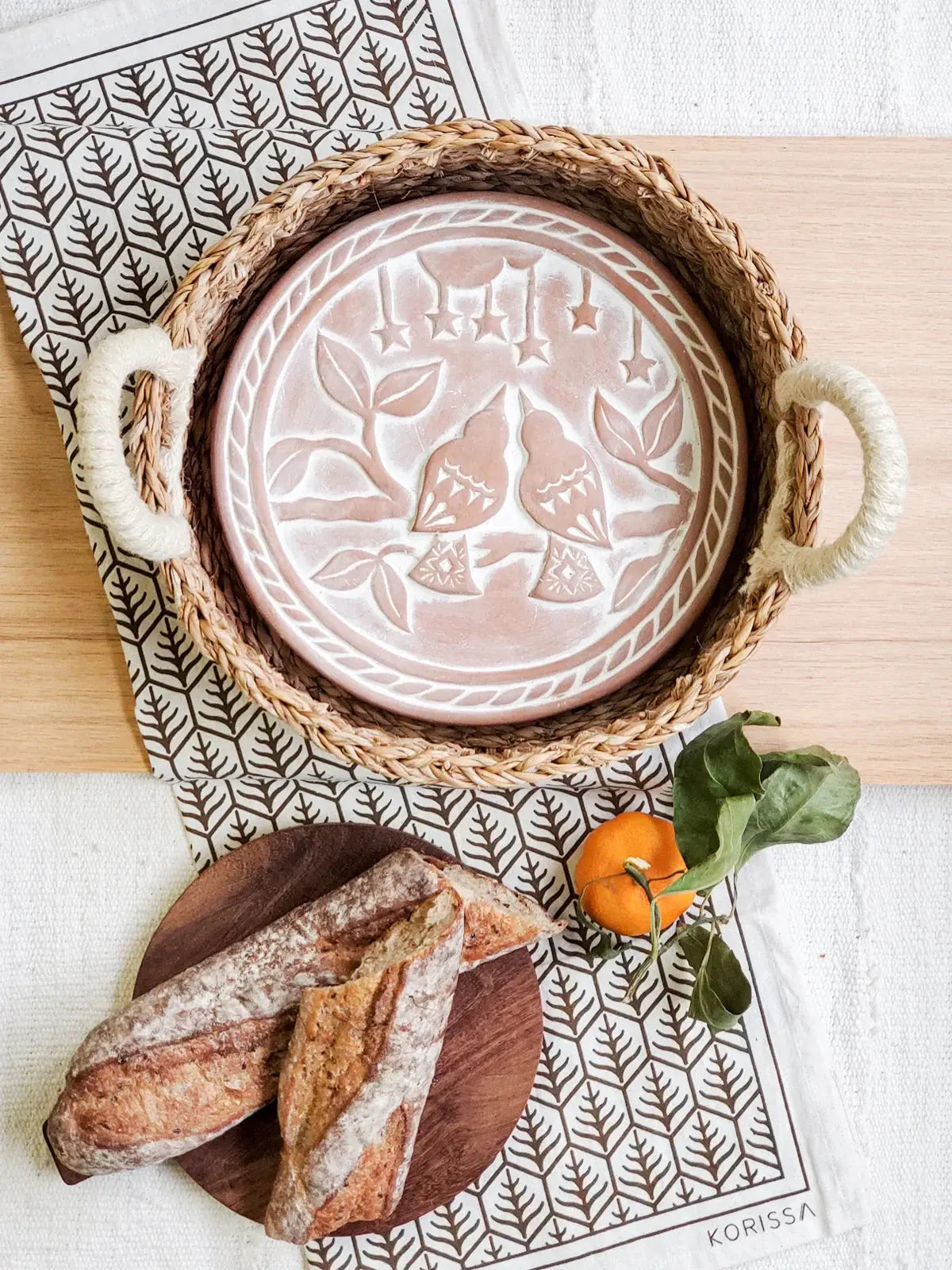 Farmhouse-style ceramic plate with bird design in woven tray, rustic bread, and orange on leaf-patterned cloth