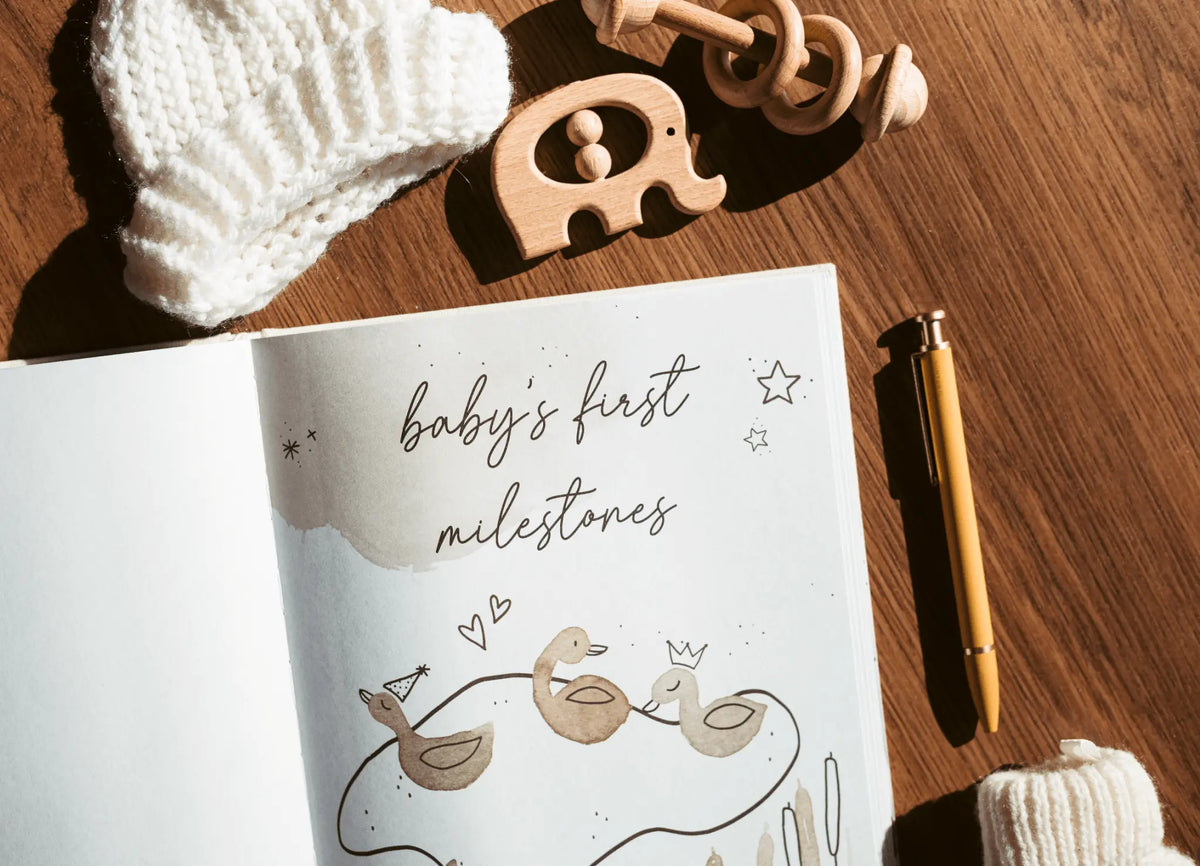 Notebook titled 'baby's first milestones' with baby items on a wooden surface