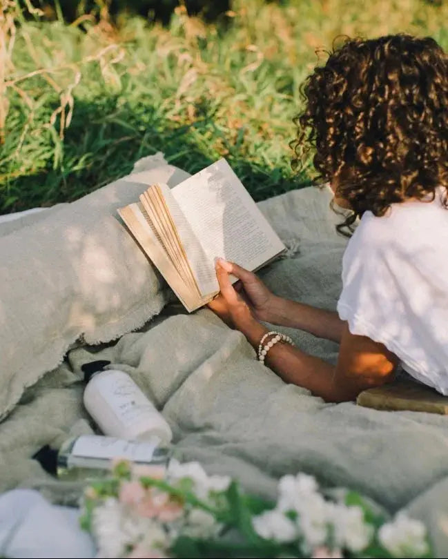 Woman reading book on cozy blanket outdoors with bath essentials and flowers, farmhouse cottage style