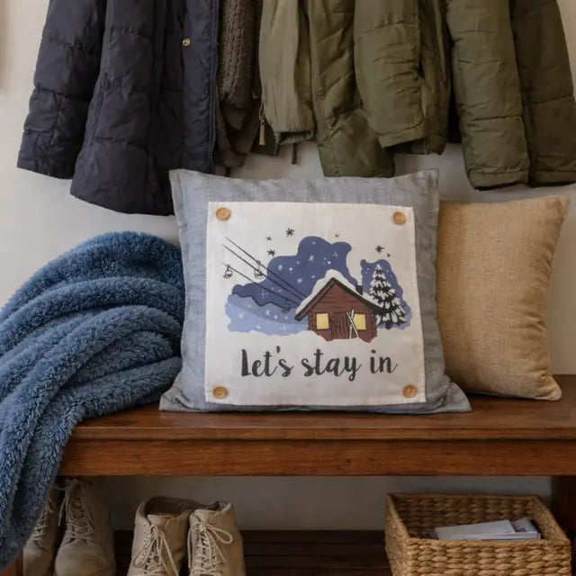 A square throw pillow with a rustic winter cabin illustration and the text 'let's stay in' sits on a wooden bench in a styled entryway. A blue blanket, a beige pillow, hanging jackets, and boots are visible around it.