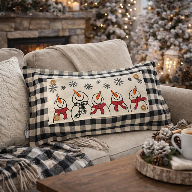 A rectangular holiday throw pillow with a black and white buffalo plaid border and a central cream panel featuring four snowmen with red scarves, orange carrot noses, and snowflake details. The pillow is styled on a beige couch in a cozy, festive living room setting with a fireplace, blanket, and holiday decorations.