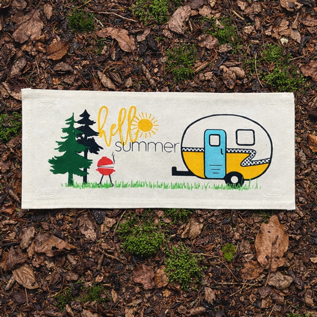 A rectangular cream-colored fabric sign or towel lies on a bed of brown leaves and moss. It features a colorful summer-themed illustration with pine trees, a red grill, yellow 'hello summer' text, and a retro trailer.