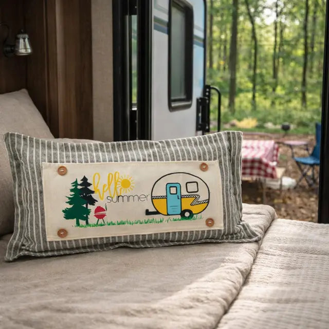 A rectangular throw pillow with a gray and white striped border and a central cream patch featuring the words 'hello summer' and camping illustrations, resting on a bed in a cozy bedroom setting.