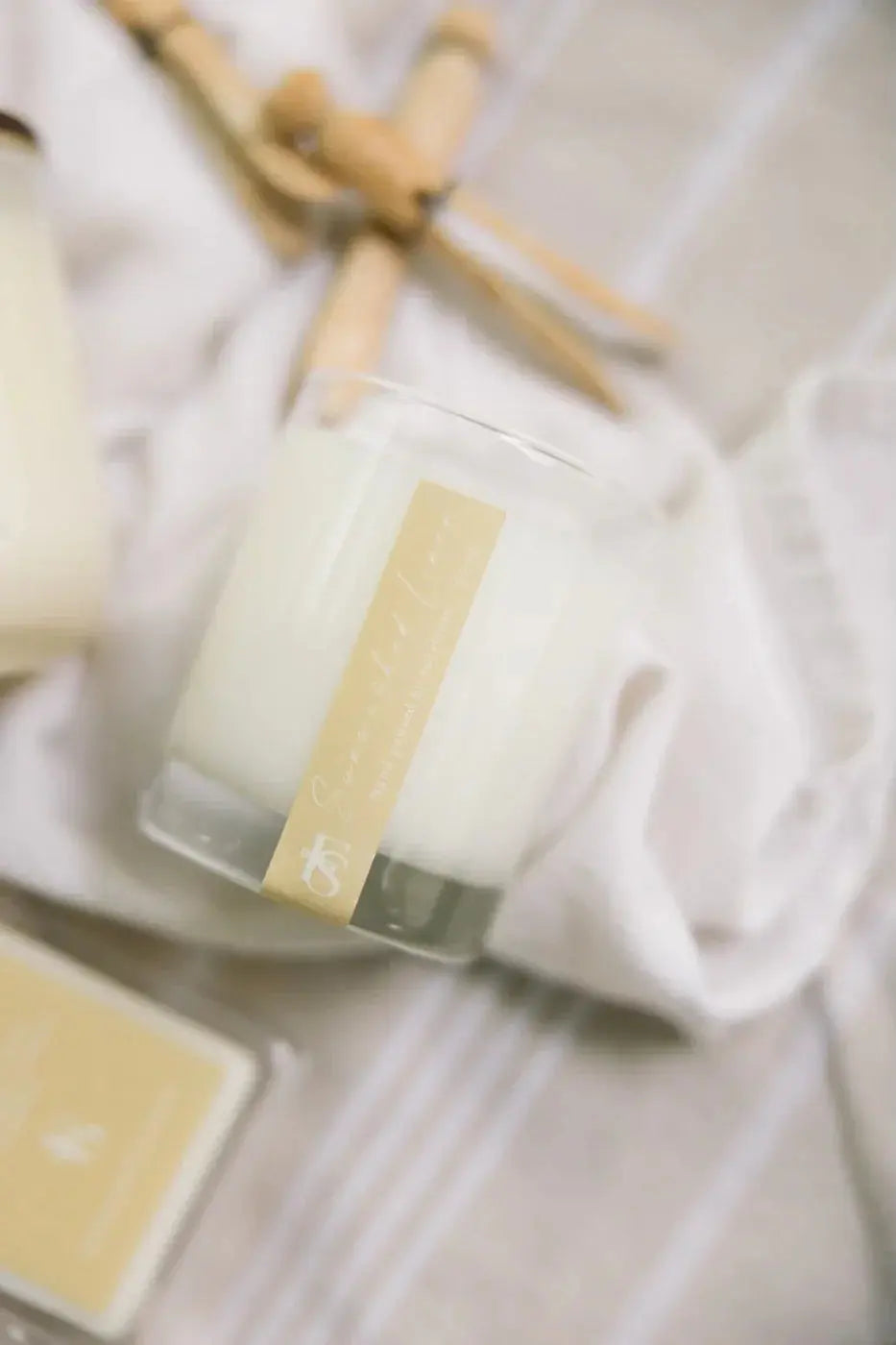 Cozy farmhouse candle in glass jar with gold label, styled on soft linens for warm home decor
