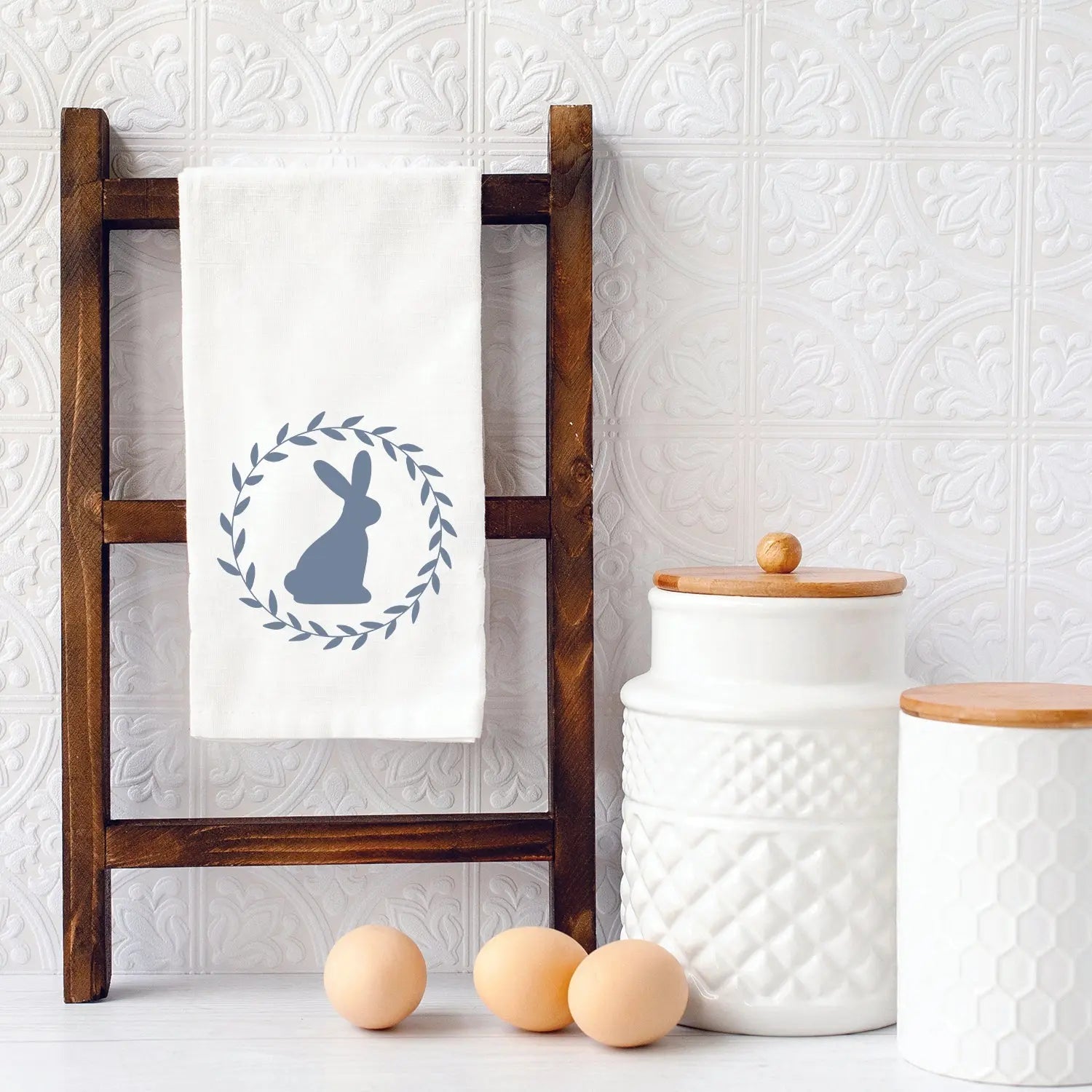 White kitchen towel with blue bunny wreath design, farmhouse decor, wooden ladder, ceramic canisters, eggs